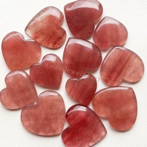 May include: A collection of twelve polished, heart-shaped strawberry quartz gemstones in varying shades of translucent reddish-pink. Each stone features a smooth, rounded surface with natural internal inclusions. The text JolariGemsJewelsEtsy.Com is visible.