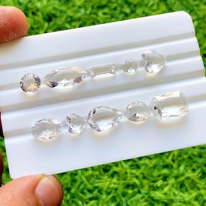 Puede incluir: Una colección de gemas transparentes de varias formas, incluyendo ovaladas, rectangulares, de corazón y de pera. Las gemas se exhiben sobre una superficie blanca y estriada, con un fondo de hierba verde. Las gemas son transparentes y reflejan la luz.