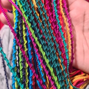 May include: A collection of multiple thin, braided cord bracelets in a vibrant array of colours including magenta, lime green, bright blue, yellow, orange, and dark navy. Each cord features a consistent, repeating twisted knot pattern throughout.