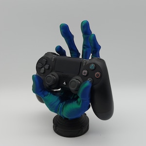Könnte beinhalten: Eine 3D-gedruckte Hand, die in blauen und grünen, schillernden Farben bemalt ist, hält einen schwarzen PlayStation 4-Controller. Die Hand steht auf einem schwarzen Sockel.