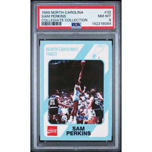 May include: A collectible trading card featuring Sam Perkins from the 1989 North Carolina Collegiate Collection. The card shows Perkins in action, reaching for a basketball, with other players in the background. The card is encased in a protective holder.