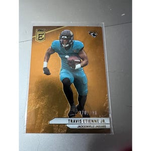 May include: A football trading card featuring Travis Etienne Jr. of the Jacksonville Jaguars. The card has a gold background with an image of the player in his teal uniform. The card is numbered 270/299.