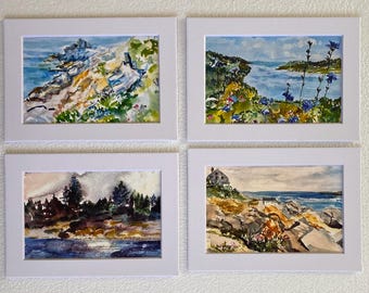 May include: Four framed watercolor paintings depicting coastal scenes. The top left painting shows a rocky coastline with blue water and green foliage. The top right painting shows a view of a bay with blue water and green hills. The bottom left painting shows a forest with a body of water in the foreground. The bottom right painting shows a rocky coastline with a house in the distance.
