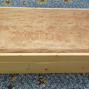 May include: A rectangular wooden box, featuring a light-coloured frame and a top made of patterned wood. The box is designed for storage or display, highlighting the natural wood grain.