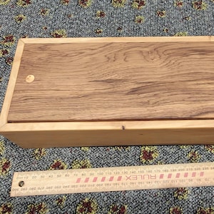 May include: A rectangular wooden box with a light-coloured frame and a darker wood grain lid. A playing card with an ace of hearts and a ruler measuring up to 300 millimetres are placed next to the box to provide a sense of scale for the object.