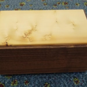 May include: A rectangular wooden box. The lid is light-coloured with a natural wood grain, contrasting with the darker brown base. The box is likely handcrafted and suitable for storage or display purposes.