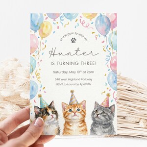 May include: A birthday invitation with a watercolour design, featuring three kittens in party hats and balloons. The text reads "Hunter IS TURNING THREE!" The invitation is held by a hand, with confetti and the words "Come paw-ty with us!"