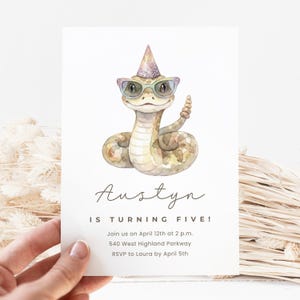 Puede incluir: Una invitación de cumpleaños con una ilustración en acuarela de una serpiente con gafas y un gorro de fiesta. El texto dice "¡Austyn cumple cinco años!" con los detalles del evento. La invitación es sostenida por una mano.