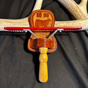 Puede incluir: Un peine de aseo rojo y plateado con un mango de madera está unido a un soporte de cuero marrón con las iniciales "HM" y una silueta de vaca.