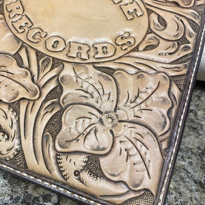 Puede incluir: Primer plano de un diario de cuero marr&oacute;n claro con un dise&ntilde;o estampado de flores y las palabras "Cathe Records" en un c&iacute;rculo. El diario tiene un borde cosido marr&oacute;n.