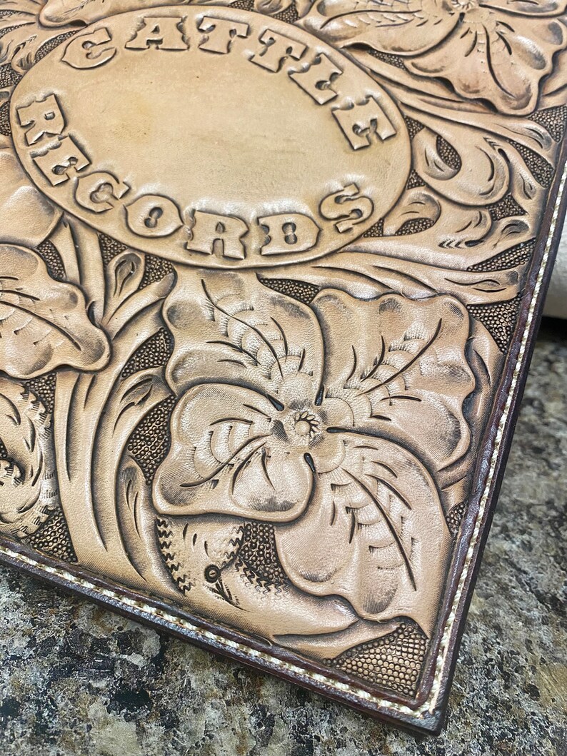 Puede incluir: Primer plano de un diario de cuero marr&oacute;n claro con un dise&ntilde;o estampado de flores y las palabras "Cathe Records" en un c&iacute;rculo. El diario tiene un borde cosido marr&oacute;n.