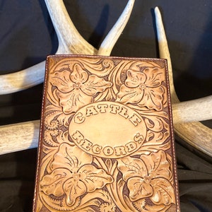 Puede incluir: Un diario de cuero marr&oacute;n con dise&ntilde;os florales tallados y las palabras "Cattle Records" en relieve en el centro.