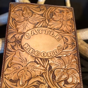 Puede incluir: Un diario de cuero marr&oacute;n con dise&ntilde;os florales tallados y las palabras "Cattle Records" en relieve en un c&iacute;rculo en la portada.