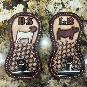 May include: Two brown leather show stick holders with a woven pattern. The left holder has a white goat silhouette and the initials "BS". The right holder has a brown pig silhouette and the initials "LB".