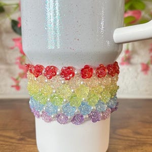 May include: White tumbler with a rainbow floral design. The tumbler has a white handle and a decorative band of colorful flowers and faux pearls. The flowers are red, yellow, green, blue, and purple.