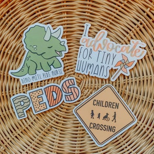 May include: Four colorful stickers with different designs. One sticker features a green dinosaur with the text "Dino-mite Peds Nurse". Another sticker features the text "Peds" in a colorful, striped font. A third sticker features a yellow and orange sign with the text "Children Crossing" and a graphic of children walking. The final sticker features the text "I advocate for tiny humans" with a pink and orange pinwheel.