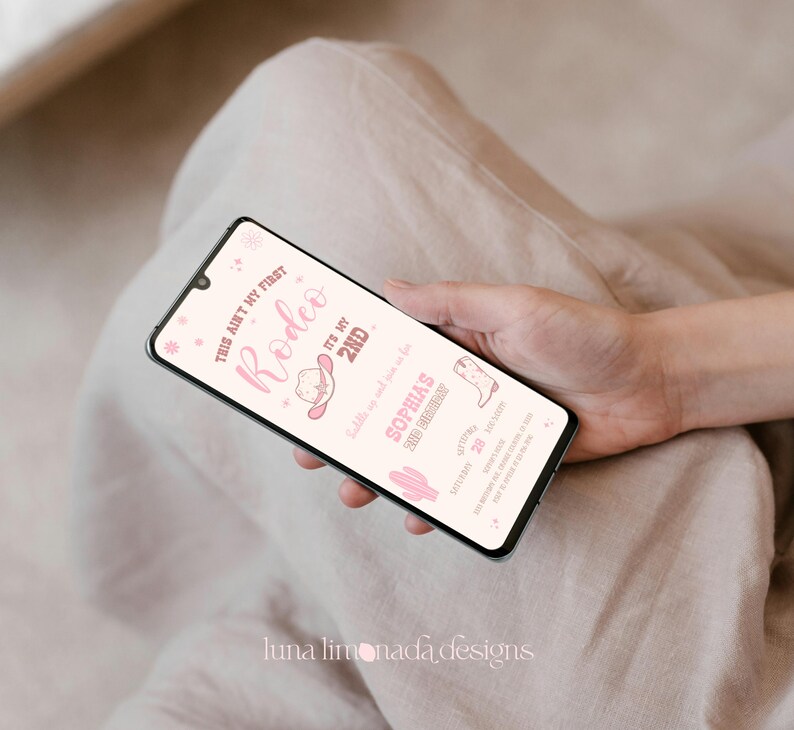 K&ouml;nnte beinhalten: Ein Smartphone mit einer digitalen Geburtstagseinladung in Rosa und Wei&szlig; mit dem Text "THIS AIN'T MY FIRST RODEO IT'S MY 2ND", "SADDLE UP AND JOIN US FOR SOPHIA'S 2ND BIRTHDAY", "SEPTEMBER 28", "12:00-3:00PM" und "123 ANYWHERE AVE, ANYWHERE, ST 12345".