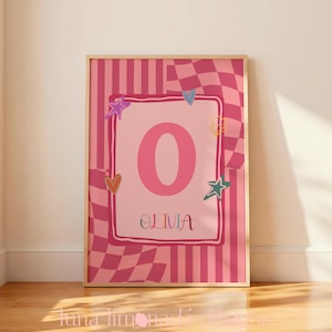 Puede incluir: Una lámina decorativa rectangular en un marco de madera clara que presenta una letra O grande de color rosa y el nombre OLIVIA en un recuadro central. El fondo muestra un patrón de cuadros y rayas en rosa y rosa oscuro con estrellas.