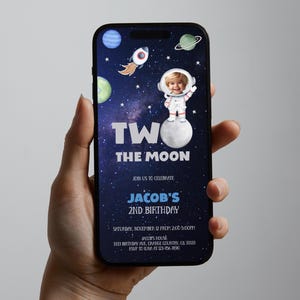 Puede incluir: Un smartphone muestra una invitación de cumpleaños con temática espacial. El diseño presenta un astronauta, planetas y el texto "Two the Moon". La invitación es para el 2º cumpleaños de Jacob, con fecha, hora y lugar.