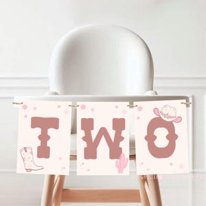 May include: Pink and brown high chair banner with the word "TWO" in large letters. The banner features a pink cowboy hat, a cactus, and a pink cowboy boot.