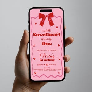 May include: Pink and red digital invitation for a first birthday party. The invitation features a red bow, hearts, and the text "our little Sweetheart is turning One". The invitation also includes the name "Olivia's 1st birthday", the date "February 23", the time "11:00 AM", the location "Luna Cafe", and the address "3333 Celebration Ave, Orange Country, CA 33333".