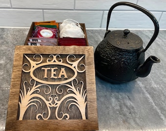 May include: A wooden tea box with an intricate design and the word "TEA" carved into the front. The box is filled with tea bags and tea sachets. A black cast iron teapot sits on a white countertop.