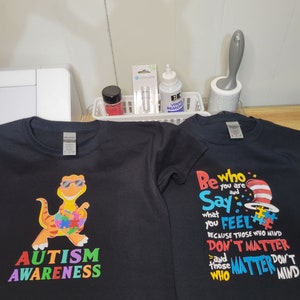 May include: Two black t-shirts with colorful designs. The first shirt features a cartoon dinosaur with a puzzle piece heart and the text "AUTISM AWARENESS". The second shirt features a red and white hat with the text "Be who you are Say what you FEEL because those who mind DON'T MATTER and those who MATTER DON'T MIND".