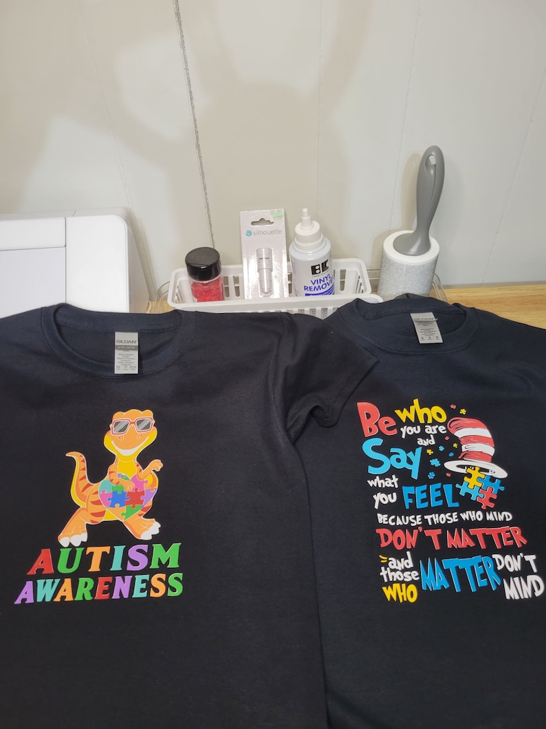 May include: Two black t-shirts with colorful designs. The first shirt features a cartoon dinosaur with a puzzle piece heart and the text "AUTISM AWARENESS". The second shirt features a red and white hat with the text "Be who you are Say what you FEEL because those who mind DON'T MATTER and those who MATTER DON'T MIND".