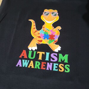 May include: Black t-shirt with a colorful dinosaur wearing sunglasses and holding a puzzle piece heart. The text "AUTISM AWARENESS" is printed below the dinosaur in rainbow colors.
