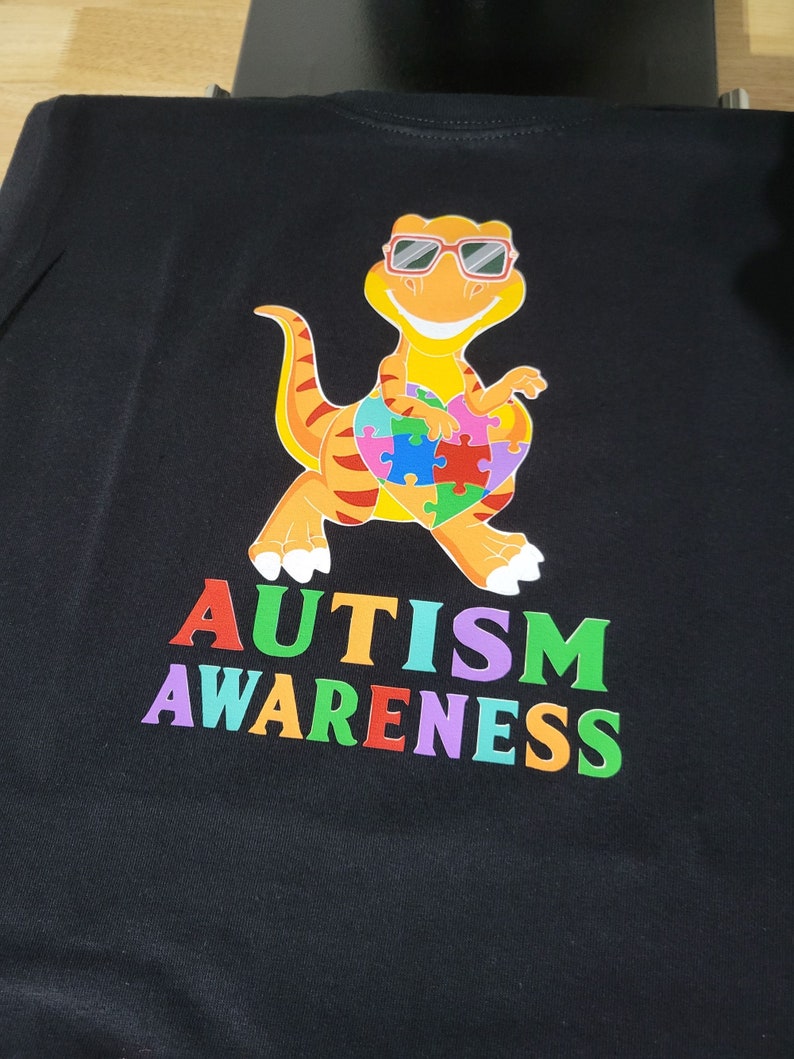 May include: Black t-shirt with a colorful dinosaur wearing sunglasses and holding a puzzle piece heart. The text "AUTISM AWARENESS" is printed below the dinosaur in rainbow colors.