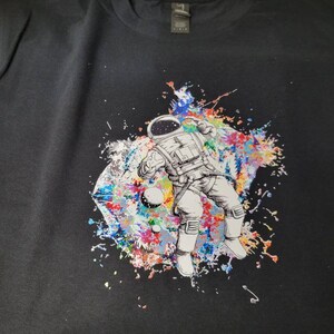 May include: Black t-shirt with a colorful graphic of an astronaut floating in space. The astronaut is surrounded by colorful paint splatters and planets.