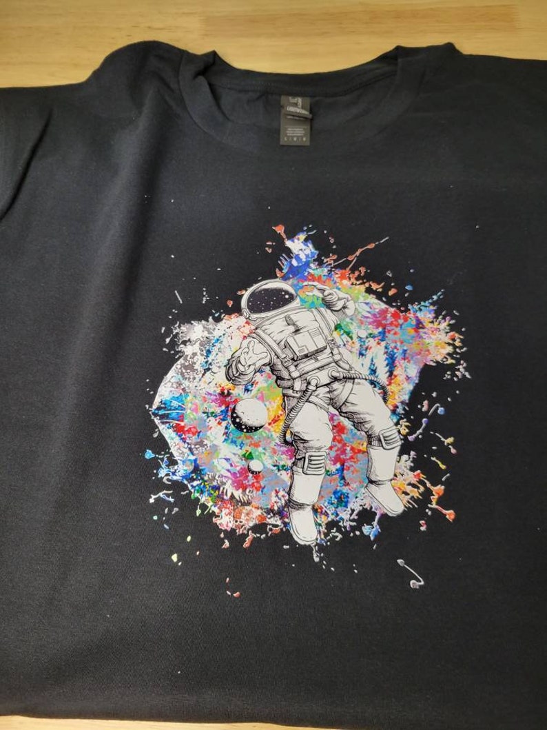 May include: Black t-shirt with a colorful graphic of an astronaut floating in space. The astronaut is surrounded by colorful paint splatters and planets.