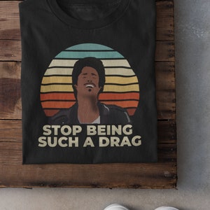 May include: Black t-shirt with a retro-style graphic of a man with an afro, a sunset, and the text "Stop Being Such A Drag".
