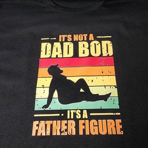 May include: Black t-shirt with a vintage-style graphic that says "It's not a dad bod. It's a father figure." The graphic features a silhouette of a person with a sunset background.
