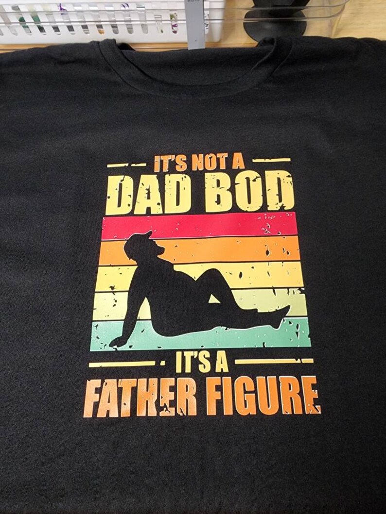 May include: Black t-shirt with a vintage-style graphic that says "It's not a dad bod. It's a father figure." The graphic features a silhouette of a person with a sunset background.