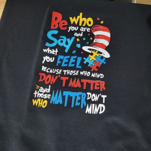 May include: Black t-shirt with a colorful graphic design featuring the text "Be who you are and Say what you FEEL because those who mind DON'T MATTER and those who MATTER DON'T MIND". The graphic also includes a red and white hat with a red and white stripe.