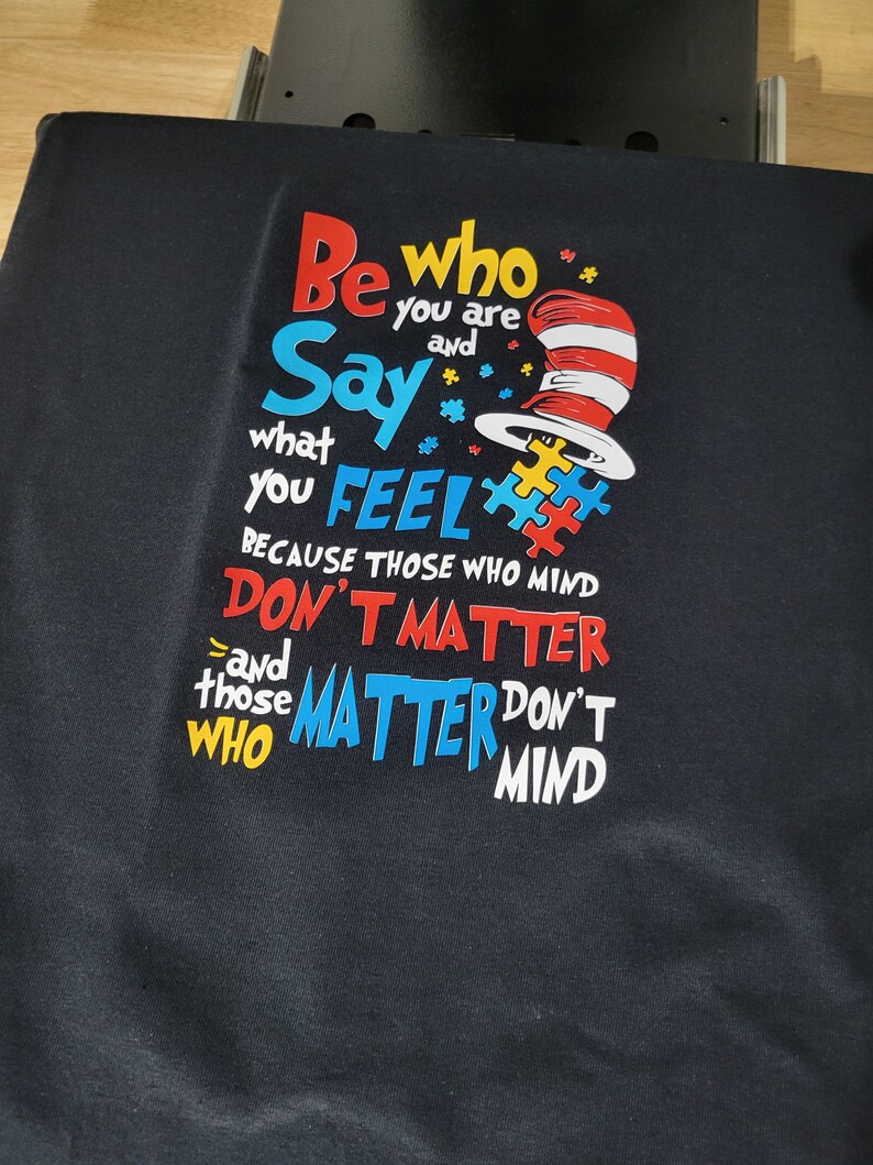 May include: Black t-shirt with a colorful graphic design featuring the text "Be who you are and Say what you FEEL because those who mind DON'T MATTER and those who MATTER DON'T MIND". The graphic also includes a red and white hat with a red and white stripe.