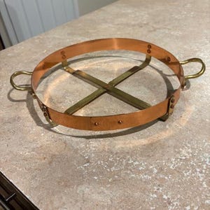 May include: A copper and brass trivet with a circular design. The trivet has a copper ring with brass handles and is supported by a brass cross-shaped base. The metal has a warm, reddish-brown hue.