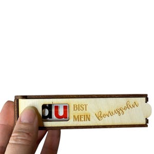 Könnte beinhalten: Eine kleine, rechteckige Holzbox mit den Worten "du BIST MEIN Bonussohn" eingeätzt. Die Box hat einen Schiebedeckel und wird von einer Hand gehalten.