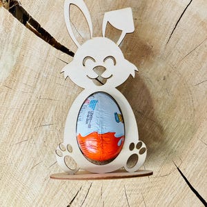 Peut inclure: Décoration en bois en forme de lapin de Pâques souriant, tenant un œuf Kinder Surprise. Le lapin est en bois clair avec des détails découpés pour les yeux, le nez et les pattes. L'œuf est enveloppé dans du papier d'aluminium orange et bleu.
