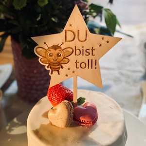 Peut inclure: Un petit gâteau blanc décoré de fraises et de macarons en forme de cœur. Un décor de gâteau en forme d'étoile en bois représente une abeille de dessin animé et le texte allemand "DU bist toll!" signifiant "Tu es génial !"
