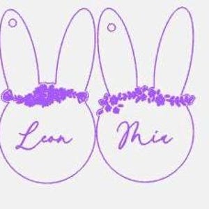 Könnte beinhalten: Zwei lila Hasen-förmige Anhänger mit Blumengirlanden und den Namen Leon und Mia in Kursivschrift.