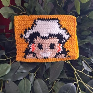May include: A handmade, crocheted wallet in a vibrant yellow color. The front features a pixelated design of a person with black hair, a white hat, and orange accents. The wallet is set against a backdrop of green leaves.