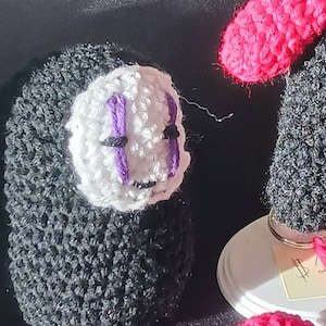 May include: A black and white crocheted amigurumi doll with a purple face and black eyes. The doll is sitting on a black surface.