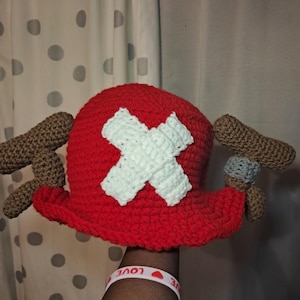 K&ouml;nnte beinhalten: Ein handgeh&auml;kelter Hut im Stil von Tony Tony Chopper aus dem Anime One Piece. Der Hut ist haupts&auml;chlich rot mit einem wei&szlig;en Kreuz auf der Vorderseite und braunen und grauen Akzenten. Ein lustiges Accessoire f&uuml;r Cosplay oder Fans.