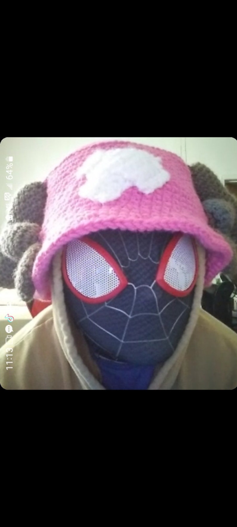 K&ouml;nnte beinhalten: Ein rosa geh&auml;kelter Hut mit einem wei&szlig;en Wolkenmuster und grauen, ohrenartigen Akzenten wird &uuml;ber einer schwarz-blauen Spider-Man-Maske getragen. Die Person tr&auml;gt einen beigen Kapuzenpullover.
