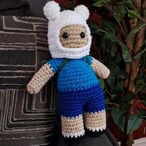 May include: A handmade crochet doll of a cartoon character, featuring a white hat with ears, a tan face, and black eyes. The doll wears a blue shirt and shorts, with tan arms and legs. The doll is approximately 10 inches tall.