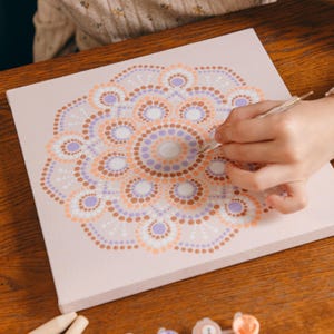May include: A canvas with a mandala design in progress. The design features concentric circles of dots in shades of peach, lavender, brown, and white. A hand is using a stylus to apply paint to the canvas, creating a dot art pattern.