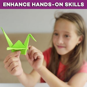 May include: A young person is holding a green origami crane. The crane is made of paper and is folded into the shape of a bird. The person is smiling and looking at the crane.