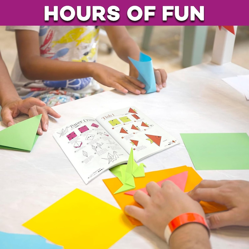 May include: Two people are folding origami paper. They are using a book with instructions for folding a paper crane and a fish. The book is open to the fish instructions. There are several pieces of origami paper in different colors on the table.